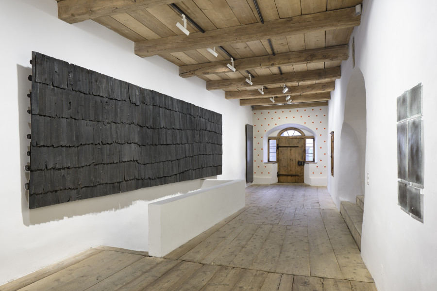 Exhibition view, Augustas Serapinas, Roof of a House from Steponių Village, 2022, charred reclaimed wooden shingle roof, 185 x 625 x 24 cm, Window Glasses from a House from Skirgiškės or Didžioji Kuosinė, 2022, window glasses with old nails powder, 90 x 59 x 0.5 cm. Photo: Ralph Feiner, Courtesy of the artist and Galerie Tschudi