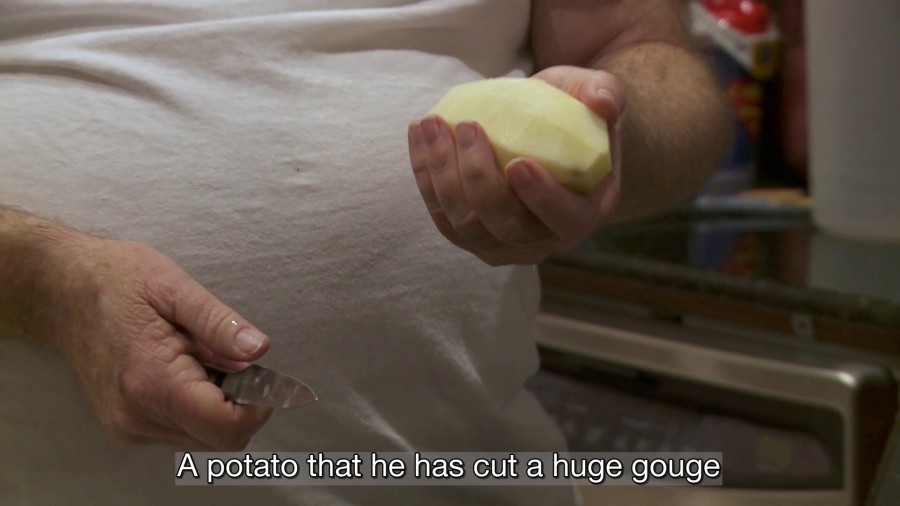 Jordan Lord, I Can Hear My Mother’s Voice, with Deborah Lord, 2018. Image description: An older white man holds a peeled potato in one hand and a knife in the other, wearing a plain white t-shirt. A caption over the image reads: “A potato that he has cut a huge gouge”.